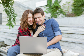 Young cheerful couple of lovers spend time together while use laptop outdoors. Happy, funny people. Love concept.