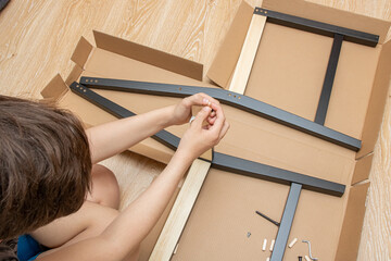 The teenager is sitting on the floor and is assembling a new black wooden chair.