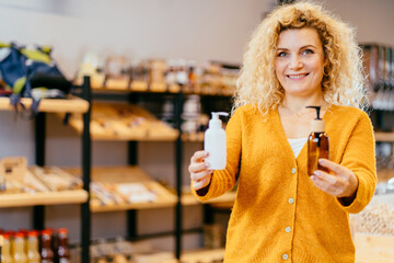 Blond happy woman choosing between plastic or glass bottle with cosmetic care product.Zero waste concept. Green and conscious lifestyle concept. on interior background of shop.sustainable lifestyle.