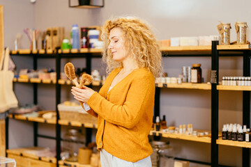 Vivacious blond curly woman on yellow background holds dishwashing brushes made from coconut flakes with bamboo handle. Biodegradable kitchen accessories. Zero waste and no plastic concept.
