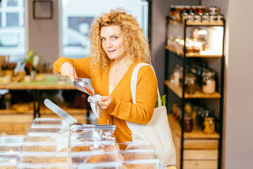 Young blond woman in yellow sweater with reusable cotton bag doing shopping in plastic free store. Minimalist vegan girl buying Goji berries, groceries without plastic packaging in zero waste shop.