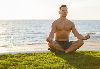 Young muscular man with naked torso meditates in lotus pose at sunrise on beach