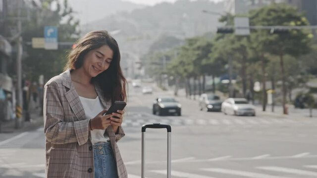 Young businesslady standing on sidewalk on street, looking at phone. Woman using smartphone, chatting during business trip. Girl using gadgets to communicate. Concept of travel, lifestyle.