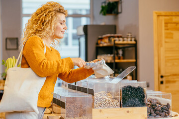 Blond girl in yellow cardigan with cotton bag puts makes conscious shopping in zero waste shop. Woman buying food, nuts in plastic free grocery store. Sustainable small business. Minimalist lifestyle.