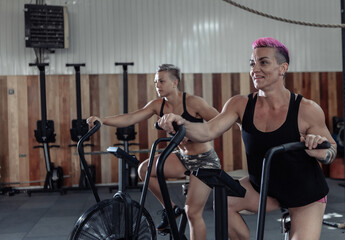 Fototapeta na wymiar Two muscular athletic women exercising on air bikes at the gym. Functional training cross class working out