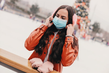 Beautiful young woman in pink earmuffs wear protective face mask, skate on outdoor ice rink. Happy stylish girl put on medical face mask on ice skating rink. Personal safety, winter holidays activity