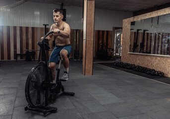 Obraz na płótnie Canvas Powerful muscular male athlete with a naked torso trains with an air bike in a modern gym. Functional, cross-fit training. Cardio exercise. Healthy lifestyle concept