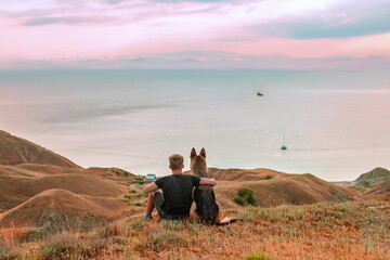 young man with his dog is sitting in the mountains and looking at the sea sunset. german shepherd...