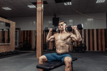 Fototapeta na wymiar Powerful athletic man training shoulder muscles with heavy dumbbells while sitting on bench in gym. Healthy lifestyle concept
