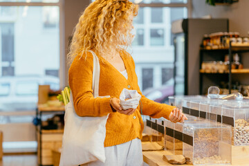 Blond curly girl in yellow cardigan with cotton bag makes conscious shopping in zero waste shop. Woman buying food in plastic free grocery store. Sustainable small businesses. Minimalist lifestyle.