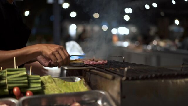 Handheld shot of man flipping over meat on sticks on the grill, at the street market food in Thailand. Close up shot of a meat steak on the grill