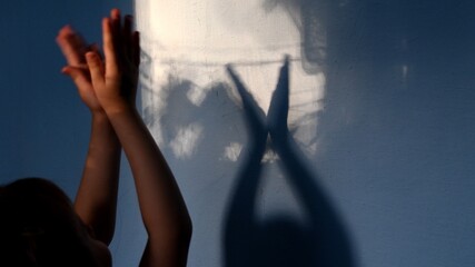 Child claps hands on unfocused foreground and focus to blurry hand shadows on wall background. Game with light and shadow. Dark spooky silhouettes of hands for Halloween concept