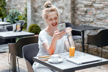 Smiling senior female blogger in casual wear sitting in coffee shop using smartphone and laptop.