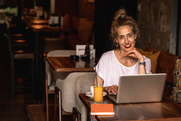 Fototapeta na wymiar Smiling senior female blogger in casual wear sitting in coffee shop using laptop.