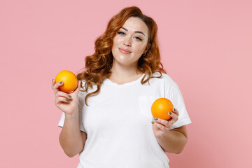 Smiling pretty young redhead plus size body positive woman 20s wearing white casual t-shirt hold ripe fresh orange fruits looking camera isolated on pastel pink color wall background studio portrait.