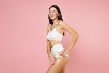 Fototapeta na wymiar Side view of cheerful beautiful thin young brunette woman 20s in white underwear posing applying moisturizer body cream from container isolated on pastel pink colour wall background, studio portrait.
