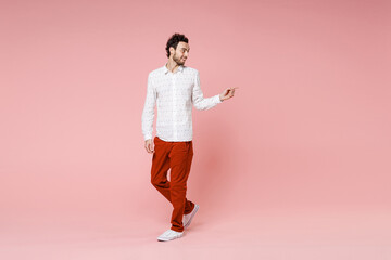 Full length of smiling young bearded man 20s wearing basic casual white shirt standing pointing index finger aside on mock up copy space isolated on pastel pink color wall background studio portrait.