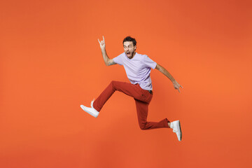 Fototapeta na wymiar Full length side view of crazy screaming young bearded man 20s in casual violet t-shirt jumping depicting heavy metal rock sign horns up gesture isolated on bright orange background studio portrait.