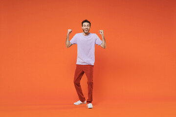 Fototapeta na wymiar Full length of overjoyed shocked young bearded man 20s in basic violet t-shirt standing doing winner gesture clenching fists looking camera isolated on bright orange wall background studio portrait.