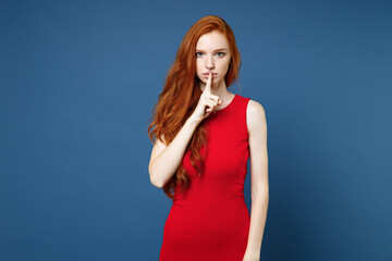 Secret young redhead woman 20s wearing bright red elegant evening dress standing saying hush be quiet with finger on lips shhh gesture looking camera isolated on blue color background studio portrait.