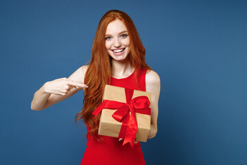 Fototapeta premium Funny young redhead woman in red elegant evening dress pointing index finger on present box with gift ribbon bow isolated on blue background. St. Valentine's Day Women's Day birthday, holiday concept.