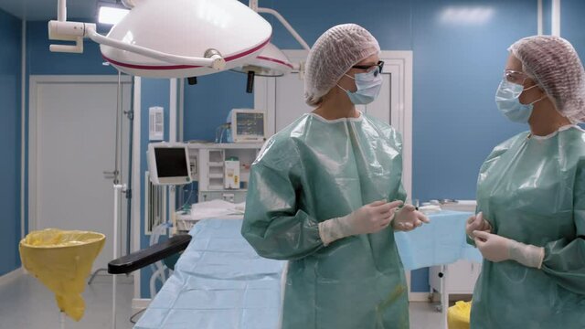 Medium close-up portrait of two female surgeons wearing sterile clothing talking in operating room before operation then looking at camera with surgical couch in background
