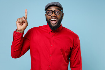 Fototapeta premium Excited young bearded african american man in casual red shirt cap eyeglasses standing holding index finger up with great new idea looking camera isolated on pastel blue background studio portrait.