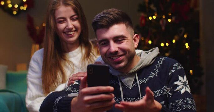 Close up of happy couple husband and wife sitting on sofa, calling on webcam, video chatting with friends using the phone, waving hand, smiling on Christmas Eve in festive mood in living room.