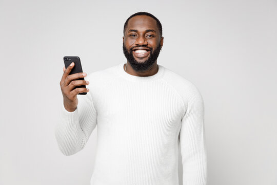 Smiling Cheerful Young African American Man 20s Wearing Casual Basic Sweater Standing Using Mobile Cell Phone Typing Sms Message Looking Camera Isolated On White Color Wall Background Studio Portrait.