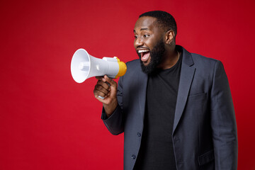 Excited cheerful funny young african american business man 20s wearing classic jacket suit standing screaming in megaphone looking aside isolated on bright red color background studio portrait.
