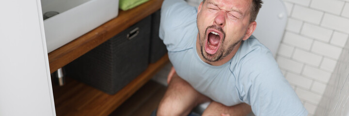 Young man with suffering on his face sits on toilet in bathroom. Indigestion constipation concept.