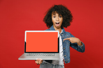 Obraz na płótnie Canvas Shocked little african american kid girl 12-13 in denim jacket pointing finger on laptop pc computer with blank empty screen isolated on red background children portrait. Childhood lifestyle concept.