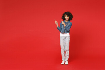 Full length excited little african american kid girl 12-13 years old in denim jacket pointing index fingers aside up isolated on red background children studio portrait. Childhood lifestyle concept.