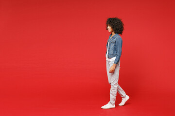 Fototapeta na wymiar Full length side view of smiling little african american kid girl 12-13 years old in casual denim jacket looking aside isolated on red background children studio portrait. Childhood lifestyle concept.
