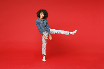 Full length of funny little african american kid girl 12-13 years old in casual denim jacket dancing rising leg isolated on bright red background children studio portrait. Childhood lifestyle concept. - obrazy, fototapety, plakaty