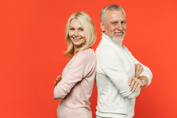 Side view of smiling couple two friends elderly gray-haired man blonde woman in white pink casual clothes standing back to back holding hands crossed isolated on orange background studio portrait.
