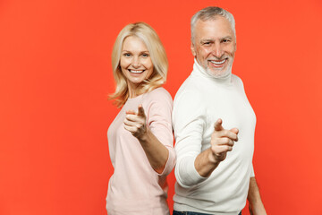 Side view of cheerful couple two friends elderly gray-haired man blonde woman in white pink clothes standing back to back pointing index finger on camera isolated on orange background studio portrait.
