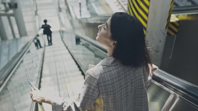 Young businesswoman going down the escalator in airport. Beautiful woman moving with baggage, looking around terminal. Girl on a way to flight for a working trip. Concept of travel, job.
