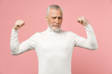 Strong handsome elderly gray-haired mustache bearded man wearing casual basic white turtleneck standing showing biceps muscles looking camera isolated on pastel pink color background studio portrait.