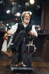 Sits on the chair in barbershop. Stylish modern senior man with gray hair and beard is indoors