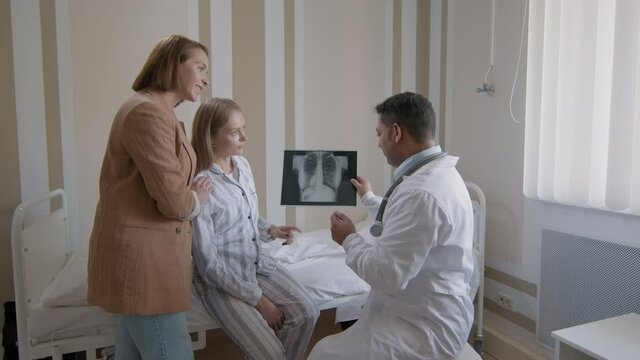 Medium footage of experienced middle-aged doctor showing picture of clean lungs on chest x-ray to young caucasian woman sitting on hospital bed