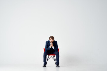business man sits on red chairs indoors and gestures with his hands on a light background