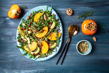 Fresh salad with fruits and greens on white wooden background vegan persimmon salad with arugula, avocado, pumpkin seeds, walnuts, pomegranate. Healthy food. top view