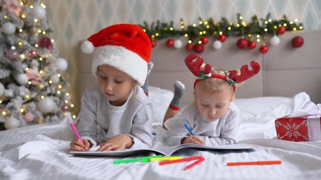 Two cute little girls sisters wearing Santa Helper hats write letter to Santa lying on bed at home in Christmas decorated room
