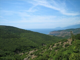 Fototapeta na wymiar A valley covered with dense low-growing forest and shrubs descends to the blue sea coast. Clouds float in the high sky.