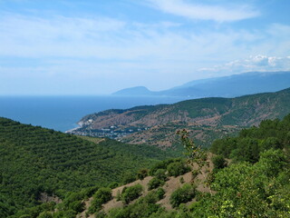 Fototapeta na wymiar A valley covered with dense low-growing forest and shrubs descends to the blue sea coast. Clouds float in the high sky.