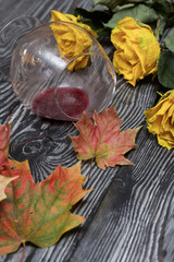 A bouquet of dried yellow roses and dried autumn leaves. An overturned glass of sour wine. On pine boards.