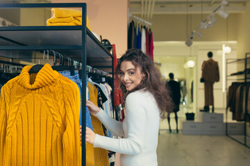 Fototapeta premium Playful attractive Female customer chooses new clothes, admiring herself in front of a mirror in a stylish boutique of shopping mall