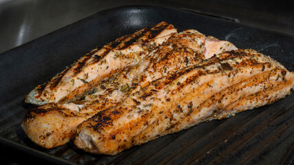Pescado cocinado dorado en plancha caliente negra hirviendo con condimento de orégano verde algunos pedazos de pimienta 