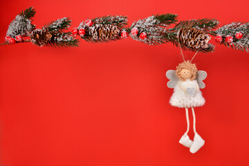 A small decorative angel girl with curly hair and a smile on her face is hanging on a Christmas tree branch with cones, covered with snow. Silver angel with a heart in her hands